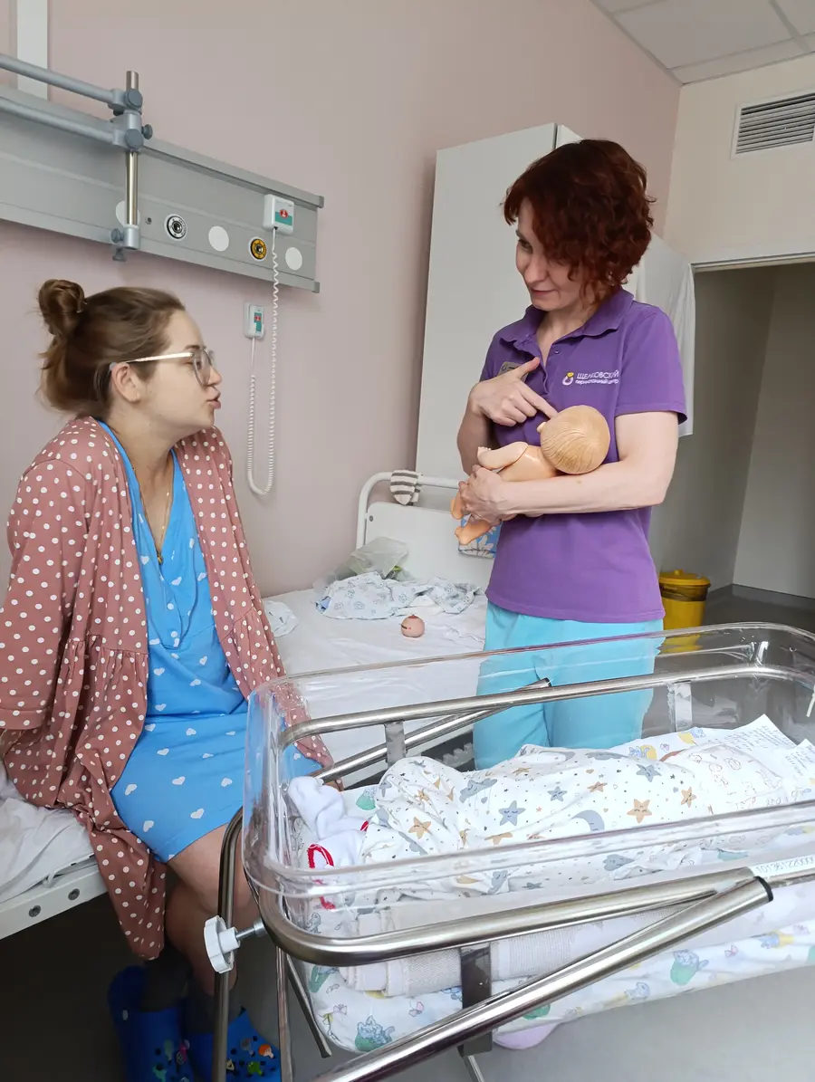 Палата роддома: женщина в халате сидит на кровати и смотрит на консультанта, которая держит у груди деревянную куклу-демонстратор и показывает положение ребёнка.